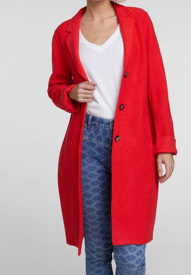 Donna che indossa un cappotto rosso brillante con bottoni sopra una maglietta bianca a scollo a V e jeans a fantasia blu, in piedi contro uno sfondo chiaro e uniforme.