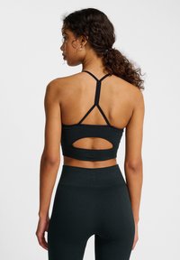 Vrouw die met haar rug naar de camera staat, gekleed in een zwarte sportieve crop top met een straps op de rug en bijpassende high-waisted leggings tegen een effen witte achtergrond.