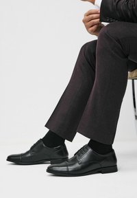 Mees istub toolil, seljas tumedad ülikondpüksid, mustad sokid ja poleeritud mustad nahast läikivad kingad.