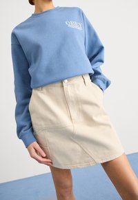 Ljusblå sweatshirt med logo och en beige denimkjol med synliga sömmar och fickor, vilket visar en avslappnad, lager-uppsättning.