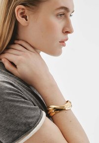 Pulsera de brazo de metal dorado con un diseño ondulado, llevada en una muñeca, sobre un fondo claro. La pulsera tiene una superficie suave y reflectante.