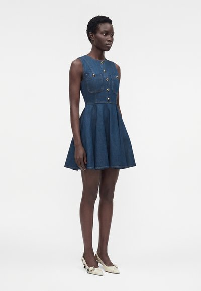 Γυναίκα στέκεται φορώντας ένα μπλε τζιν φόρεμα χωρίς μανίκια με χρυσά κουμπιά και μπροστινές τσέπες, σε συνδυασμό με λευκές μυτερές ψηλοτάκουνα.