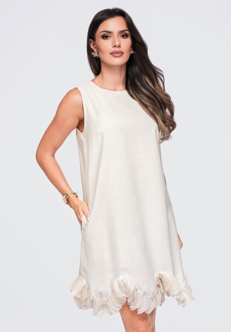 Femme aux longs cheveux foncés portant une robe crème sans manches avec un ourlet volanté et des bracelets en or, posant avec une main dans la poche.