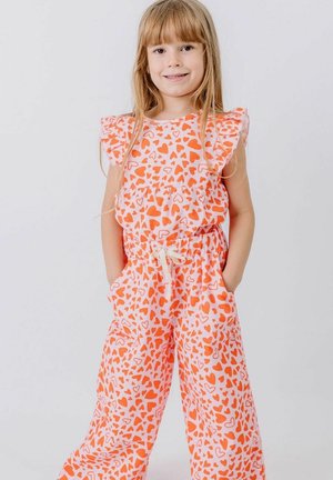 Young girl smiling with hands in pockets, wearing white jumpsuit covered in orange heart patterns and ruffled sleeves, standing on plain background.