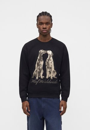 Jeune homme portant un sweat-shirt noir avec des chiens dalmatiens à la bouche ouverte se faisant face et le texte « Huf Worldwide » en dessous.