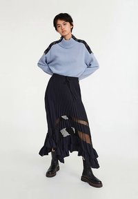 Svetlo modra pulover z visokim ovratnikom in kontrastnimi črnimi naramnicami, kombiniran z dolgo, pliseirano mornarsko krilo z nabranim robom in prozornimi poudarki.