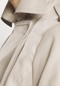 Beżowy płaszcz trencz z kołnierzem, charakteryzujący się gładką fakturą i szczegółowym przeszyciem wzdłuż szwów i kieszeni. Widoczne subtelne, klasyczne elementy designu.