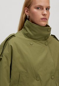 Olijfgroene trenchcoat gemaakt van een gladde stof, met een hoge kraag, dubbele knoopsluiting en een zak aan de zijkant.