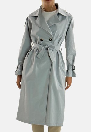 Personne portant un trench-coat gris clair ceinturé avec des boutons croisés et des poignets à boucle par-dessus un haut blanc.