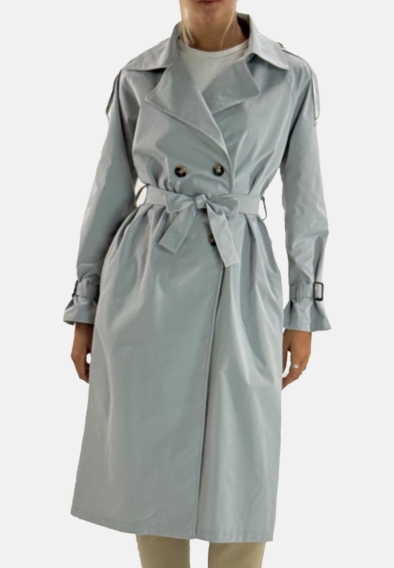 Personne portant un trench-coat gris clair ceinturé avec des boutons croisés et des poignets à boucle par-dessus un haut blanc.