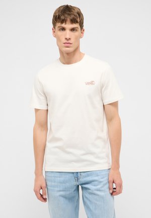 Junger Mann trägt ein schlichtes cremefarbenes T-Shirt mit kleinem Mustang-Logo und hellblauen Jeans, steht vor weißem Hintergrund.