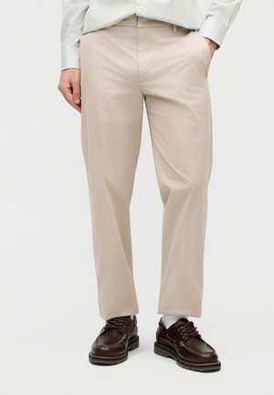 Pantalon beige droit associé à des chaussures à lacets en cuir marron foncé et des chaussettes blanches, porté avec une chemise rayée.