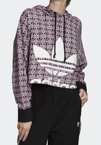 Zkrácená růžová mikina s geometrickými černými vzory, bílým logem Adidas, nastavitelnou kapucí s černými šňůrkami a černými žebrovanými manžetami.