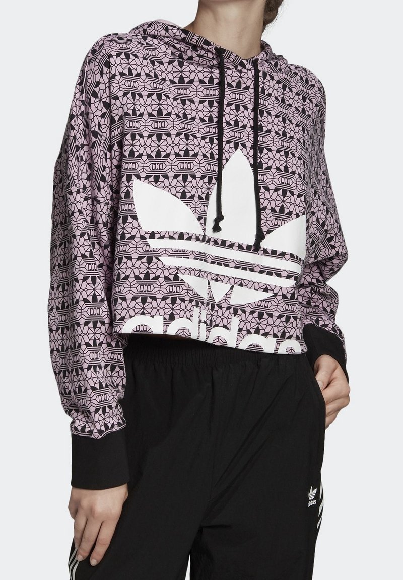 Zkrácená růžová mikina s geometrickými černými vzory, bílým logem Adidas, nastavitelnou kapucí s černými šňůrkami a černými žebrovanými manžetami.