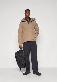 Veste imperméable beige pour hommes avec capuche, pantalon noir et sac à dos noir, dotée de poches et d'une coupe décontractée.