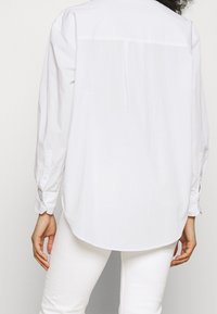Camicia bianca con bottoni, maniche lunghe, orlo arrotondato e texture liscia. Presenta bottoni ai polsini e un design semplice e minimalista.
