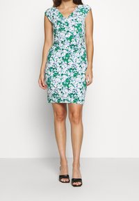 Blommig klänning med v-ringning, korta ärmar och figurnära silhuett. Tyget har gröna och blå blommönster på en ljus bakgrund.