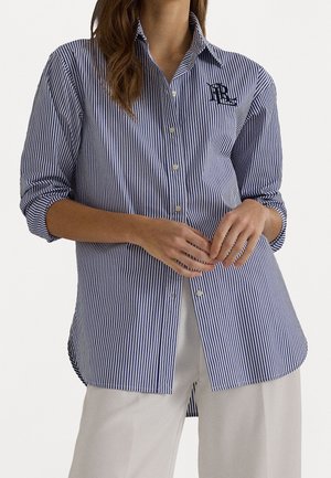 Nainen, joka pukeutunut sinivalkoraidalliseen pystybutton-up-paitaan, jossa on käännetyt hihat, ja valkoisiin housuihin, kädet rentosti vyötäröllä.