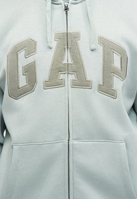 Ανοιχτό μπλε φούτερ με φερμουάρ και μεγάλες μπεζ γράμματα "GAP" στο στήθος, με κορδόνια που κρέμονται κοντά στο φερμουάρ.