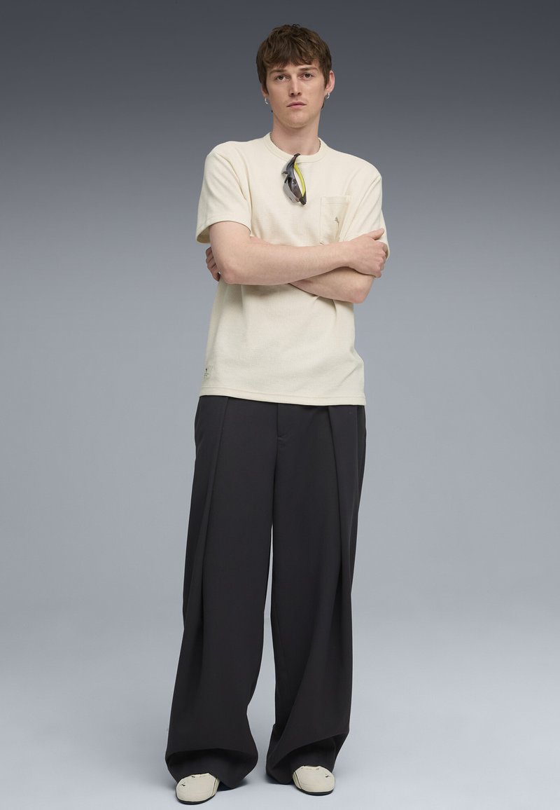 Chemise beige à manches courtes avec poche ; pantalon noir ample ; baskets blanches ; modèle avec des lunettes de soleil reposant sur la chemise, les bras croisés.