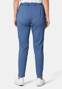 Pantalons bleus extensibles avec une taille et des plis, présentant un design à jambes fuselées. Portés avec des baskets blanches. Texture lisse.