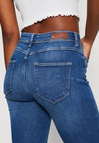 Blå denimjeans med høy midje, med to baklommer, kontrastsømmer og en brun skinnmerkelapp i midjen.