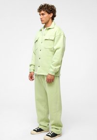 Lichtgroene, oversized jas met twee voorzakken en drukknoopsluiting, gecombineerd met bijpassende baggy broek en zwarte sneakers. Soepele textuur.