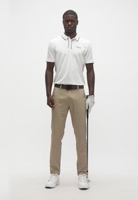 Άνδρας που στέκεται με μπεζ παντελόνι, λευκό πόλο πουκάμισο και λευκό γάντι του γκολφ, κρατώντας ένα γκολφ κλαμπ σε απλό φόντο.