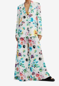 Biały żakiet w kwiaty i zestaw spodni z szerokimi nogawkami z kolorowymi kwiatami w odcieniach niebieskiego, różowego, żółtego i czarnego, noszony z czarnymi sandałami na platformie.