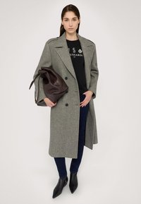 Abrigo de lana gris con diseño de doble botonadura, camiseta gráfica negra, jeans oscuros y botas de tobillo negras con punta. Sostiene un bolso de cuero marrón.