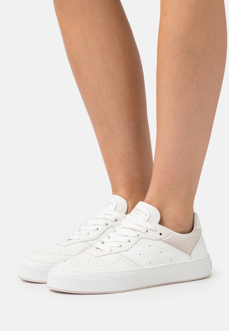 Paire de baskets blanches basses avec des détails perforés portées pieds nus, présentées sur un fond clair uni.