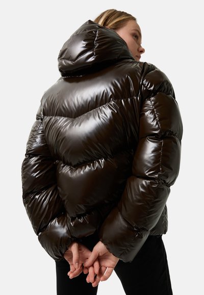Kvinde iført en skinnende mørkebrun pufferjakke med hætte, hænderne samlet bag ryggen, vendt lidt til siden mod en ensfarvet baggrund.