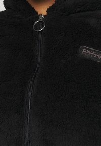 Svart fleecezipjacka med en strukturerad yta, silverfärgad dragkedja och en logopatch på vänster sida. Mjuk och varm material.