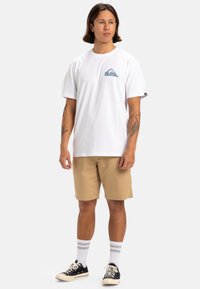 Camiseta blanca de manga corta con un gráfico de olas en el lado izquierdo del pecho, combinada con pantalones cortos beige y zapatillas negras con suelas blancas y calcetas a rayas.