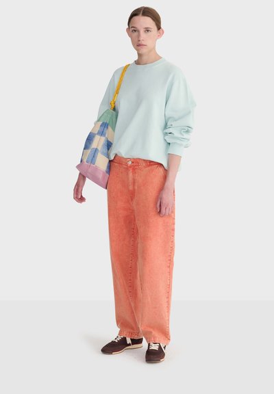 Jeune femme portant un sweat-shirt bleu clair, un pantalon ample orange, des baskets sombres et portant un sac patchwork multicolore avec une sangle jaune.