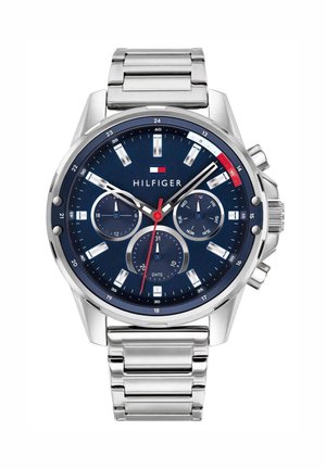 Reloj de pulsera de metal plateado con esfera azul, tres subesferas para 24 horas, día de la semana y fecha, y logotipo de Tommy Hilfiger debajo de las 12 en punto.