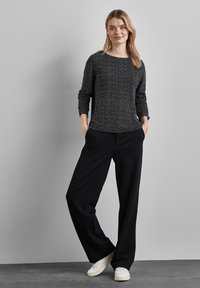 Schwarzes Langarmshirt mit einem dezenten weißen Muster, kombiniert mit lässigen schwarzen Hosen und weißen Sneakers, vor einem neutralen Hintergrund stehend.