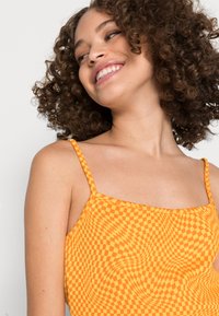Top de padrão xadrez laranja com finas alças spaghetti, apresentando um design justo e uma textura suave. O fundo é neutro e discreto.