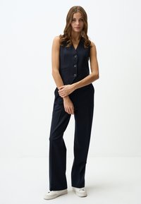 Marineblauwe jumpsuit met een mouwloos ontwerp, v-hals en een sluiting met drie knopen aan de voorkant. Tailored fit met rechte, vloeiende pijpen en zijzakken.