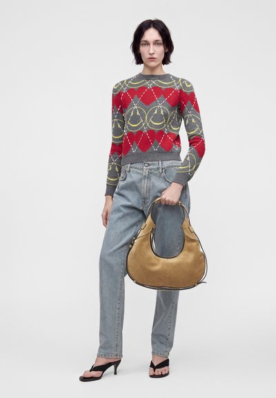 Femme portant un pull rouge et gris avec des cœurs et des visages souriants, un jean bleu clair, des sandales noires à talons, tenant un grand sac à main beige.