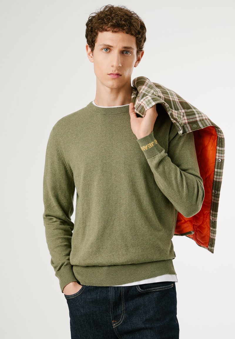 Maglione verde a maglia con polsini e orlo a coste, che presenta un logo. Il modello tiene su una spalla un giubbotto a quadri con fodera arancione.