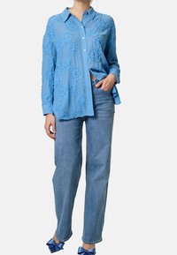 Chemise bleu clair avec des accents floraux texturés, design à boutons, manches longues et coupe décontractée. Associée à un jean droit délavé.