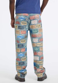 Osoba nosząca niebieskie dżinsy ozdobione kolorowymi logo Guess Originals od talii do kostek, w połączeniu z czarnymi butami.