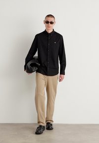 Black button-up shirt, beige trousers, holding a black jacket. Wears dark sunglasses and black shoes. Smooth fabric, simple design.