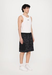 Svarte mønstrede shorts med en ensfarget svart lomme, kombinert med en hvit tanktopp som har en liten logo. Modellen har på seg hvite sneakers.
