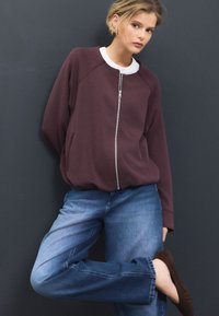 Vīnsarkanā rāvējslēdzēja džemperis ar baltu apkakli, brīva piegriezuma un sānu kabatām, kombinēts ar ziliem taisnas kājas džinsa biksēm un brūnām kurpēm ar papēžiem.