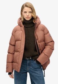 Veste matelassée brune avec capuche, présentant un design matelassé, un col montant et des accents noirs, portée sur un pull épais marron foncé et un jean bleu.