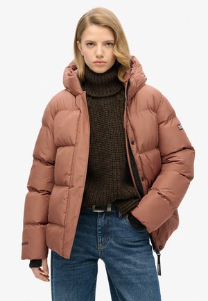 Bruine pufferjack met capuchon, met een doorgestikt ontwerp, hoge kraag en zwarte accenten, gedragen over een dikke donkerbruine trui en blauwe spijkerbroek.