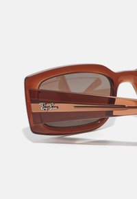 Óculos de sol retangulares castanhos com lentes castanhas transparentes, acabamentos de metal e o logótipo da Ray-Ban na armação. Superfície de plástico suave.