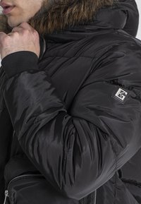 Giacca a vento nera con cappuccio foderato in pelliccia sintetica, polsini a coste e una patch con il marchio sulla manica. Presenta una tasca con zip nella parte anteriore.